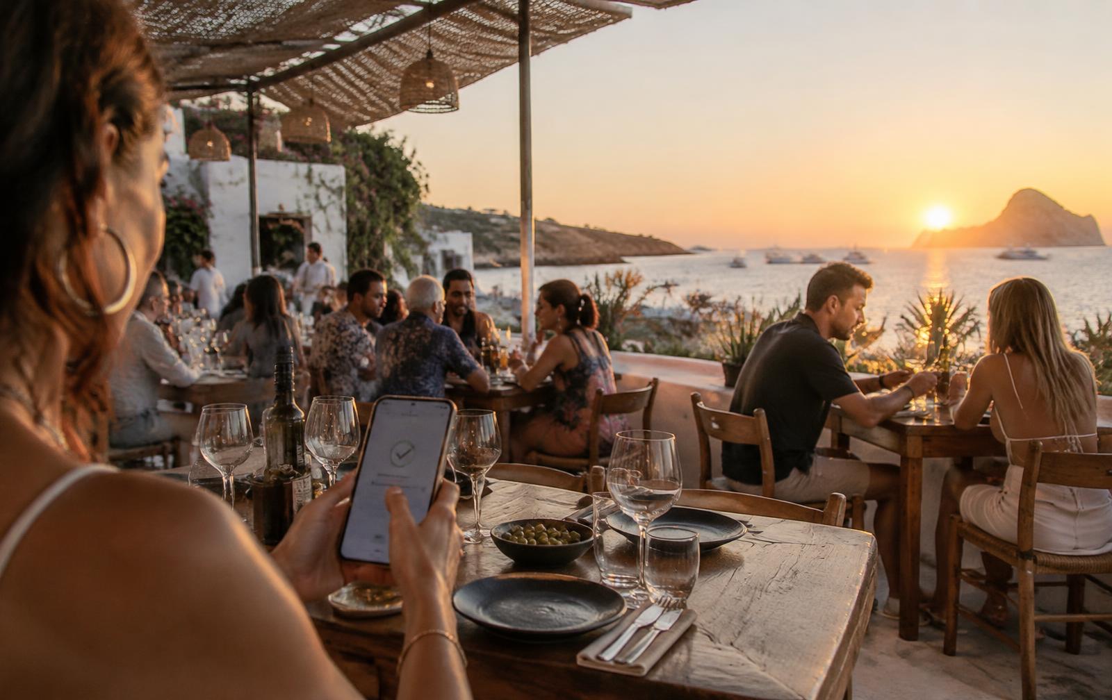 Terraza de restaurante mediterráneo en Ibiza llena de comensales al atardecer, con un móvil mostrando una reserva confirmada